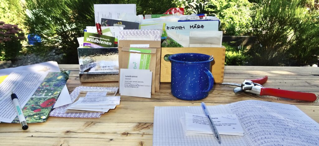 Viele Samentüten, ein Notizheft mit Kugelschreiber und eine Tasse auf einem Tisch.